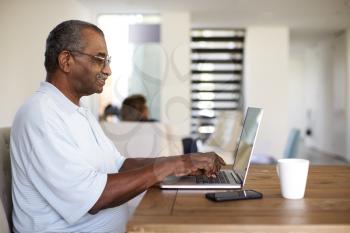 Senior black man sitting at the table using a laptop computer at home, side view