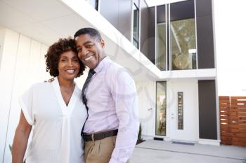 Middle aged black couple stand outside looking to camera in front of their modern home, close up