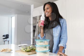 Woman In Kitchen Preparing Healthy Meal Drinking Protein Shake From Bottle