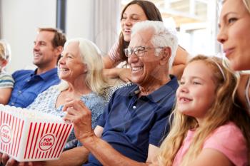 Multi-Generation Family Sitting On Sofa At Home Eating Popcorn And Watching Movie Together