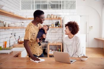 Multi-Tasking Father Holds Baby Son And Cleans Surface As Mother Uses Laptop And Eats Breakfast