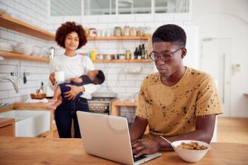 Multi-Tasking Mother Holds Baby Son And Makes Hot Drink As Father Uses Laptop And Eats Breakfast
