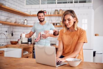 Multi-Tasking Father Holds Baby Son And Makes Hot Drink As Mother Uses Laptop And Eats Breakfast