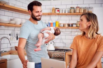 Multi-Tasking Father Holds Baby Son And Makes Hot Drink As Mother Uses Laptop And Eats Breakfast