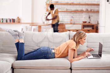 Woman Relaxing Lying On Sofa At Home Using Laptop Computer With Man In Kitchen Behind