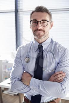 Portrait Of Smiling Mature Male Doctor With Stethoscope Standing By Desk In Office