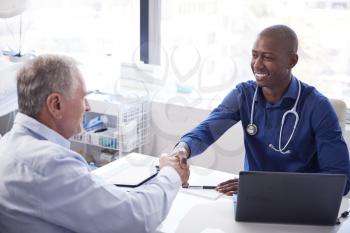 Senior Male Patient Shaking Hands With Doctor Sitting At Desk In Office