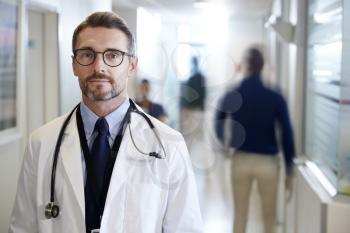 Portrait Of Mature Male Doctor Wearing White Coat With Stethoscope In Busy Hospital Corridor