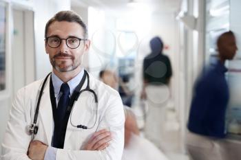 Portrait Of Mature Male Doctor Wearing White Coat With Stethoscope In Busy Hospital Corridor