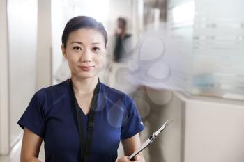 Portrait Of Female Nurse Wearing Scrubs With Clipboard In Busy Hospital Corridor