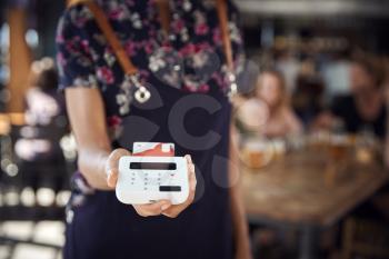 Close Up Of Waitress Holding Credit Card Payment Terminal In Busy Bar Restaurant