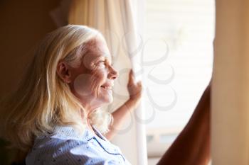 Smiling senior white woman opening the curtains on a sunny morning, side view, close up