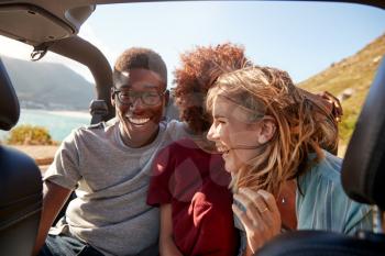 Three happy young adult friends sitting in an open car by the coast, close up, waist up