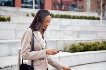 Businesswoman Commuting To Work Checking Messages On Mobile Phone Outside Modern Office Building
