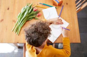 Overhead Shot Of Young Boy At Home With Bunch Of Flowers Writing In Mothers Day Card