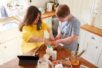Young Downs Syndrome Couple Following Recipe On Digital Tablet To Bake Cake In Kitchen At Home