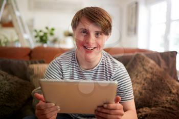 Portrait Of Young Downs Syndrome Man Sitting On Sofa Using Digital Tablet At Home