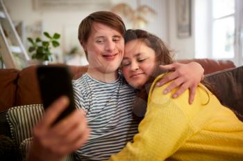 Loving Young Downs Syndrome Couple Sitting On Sofa Using Mobile Phone To Take Selfie At Home
