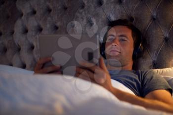 Man Wearing Wireless Headphones Lying In Bed Watching Digital Tablet Before Going To Sleep