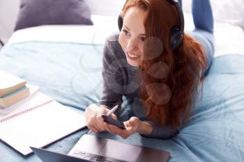 Looking Down On Female College Student Wearing Headphones Lying On Bed With Mobile Phone Working On Laptop