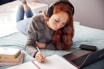 Looking Down On Female College Student Wearing Headphones Lying On Bed Working On Laptop