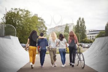 Rear View Of Female Friends With Skateboards And Bike Walking Through Urban Skate Park