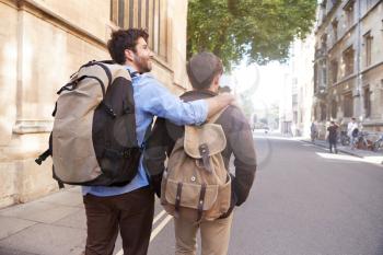 Rear View Of Male Gay Couple On Vacation Wearing Backpacks Walking Along City Street
