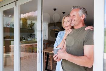 Senior Couple Standing And Looking Out Of Kitchen Door Drinking Coffee