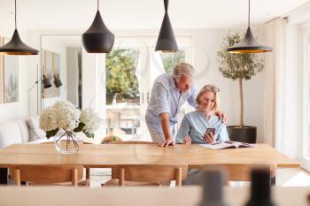 Senior Couple Relaxing With Magazine At Home Looking At Mobile Phone Sitting At Dining Room Table