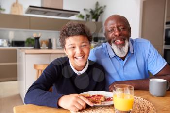 Portrait Of Smiling Grandfather Sitting In Kitchen With Grandson Eating Breakfast Before School