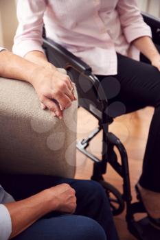 Close Up Of Mature Couple With Woman In Wheelchair Sitting In Lounge At Home Holding Hands Together