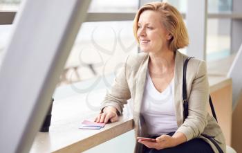 Mature Businesswoman With Passport And Boarding Pass In Airport Departure Lounge Using Mobile Phone