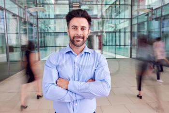 Portrait Of Businessman With Crossed Arms Standing In Lobby Of Busy Modern Office