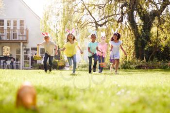 Group Of Children Wearing Bunny Ears Running To Pick Up Chocolate Egg On Easter Egg Hunt In Garden