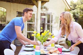 Parents Putting Food On Table For Childrens Outdoor Easter Party In Garden At Home