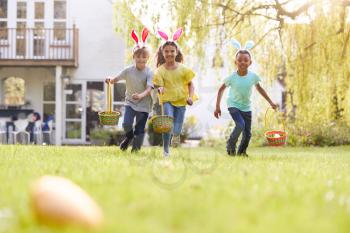 Group Of Children Wearing Bunny Ears Running On Easter Egg Hunt In Garden