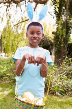 Portrait Of Boy Wearing Bunny Ears Holding Chocolate Egg On Easter Egg Hunt In Garden