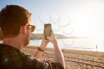 Young Man Taking Photo Of Beach In Wales On Mobile Phone