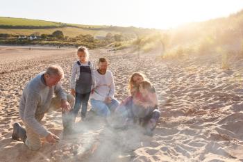 Grandfather Cooking As Multi-Generation Family Having Evening Barbecue Around Fire On Beach Vacation