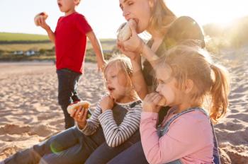 Mother With Children Eating Hot Dogs Sitting On Sand At Beach Barbecue