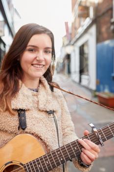 Portrait Of Young Female Musician Busking Playing Acoustic Guitar And Singing Outdoors In Street