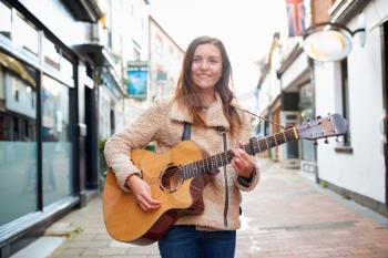 Portrait Of Young Female Musician Busking Playing Acoustic Guitar And Singing Outdoors In Street