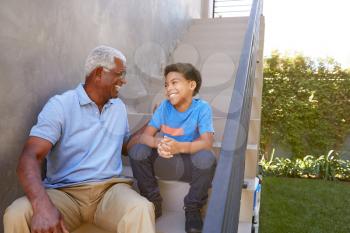 Grandfather With Grandson Sitting On Steps And Talking Outdoors At Home