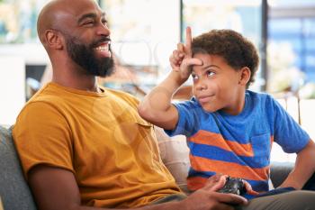 Son Makes Loser Gesture At Father As They Sit On Sofa And Play Video Game