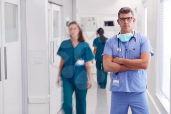 Portrait Of Mature Male Doctor Wearing Scrubs Standing In Busy Hospital Corridor