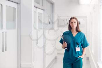 Portrait Of Female Doctor Wearing Scrubs Standing In Hospital Corridor Holding Clipboard