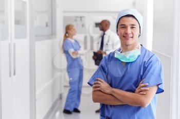 Portrait Of Male Doctor Wearing Scrubs Standing In Busy Hospital Corridor