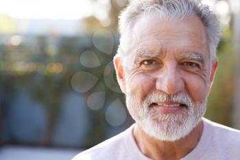 Close Up Portrait Of Smiling Retired Senior Hispanic Man In Garden At Home Against Flaring Sun