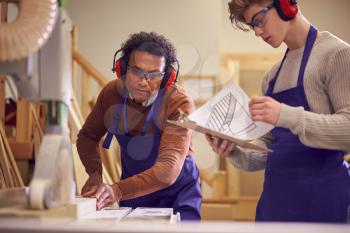 Tutor With Male Carpentry Student In Workshop Studying For Apprenticeship At College Using Bench Saw