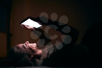 Smiling Woman With Face Illuminated By Digital Tablet Screen Lying On Carpet At Night